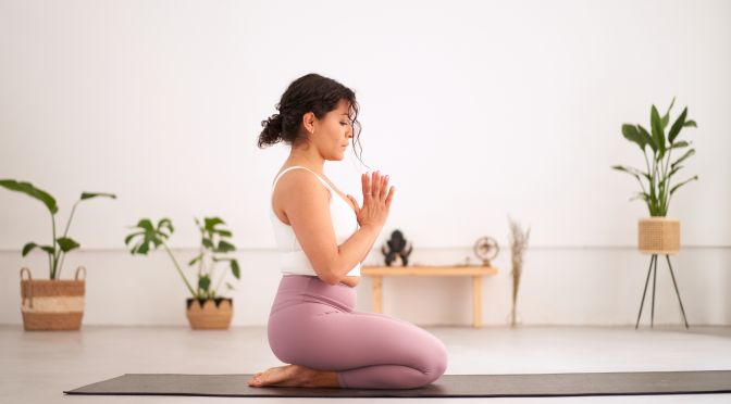 Side view of Latin woman in sportswear doing yoga exercise. Young girl concentrating performing asana anjali mudra indoors. Concept of feminine wellness, meditation, body care and mental health.